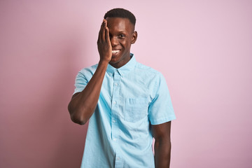 African american man wearing blue casual shirt standing over isolated pink background covering one eye with hand, confident smile on face and surprise emotion.