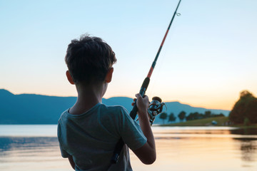 A happy boy is standing on the shore of a beautiful lake and fishing with a fishing rod against the...