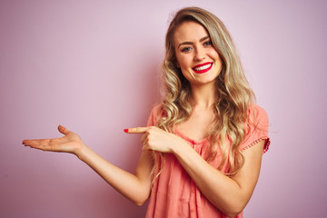 Young beautiful woman wearing t-shirt standing over pink isolated background amazed and smiling to the camera while presenting with hand and pointing with finger.