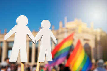 White wooden figures of gay couple holding hands with rainbow LGBT flags and Barcelona city on the background. LGBT and Pride parade concept. LGBTQI+ community
