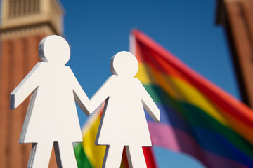 Wooden figures of two girls holding hands with rainbow LGBT flag on the background during annual Barcelona gay Pride parade. Lesbian couple. LGBT and Pride parade concept. LGBTQI+ community