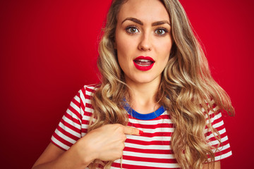 Young beautiful woman wearing stripes t-shirt standing over red isolated background with surprise face pointing finger to himself