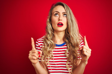 Young beautiful woman wearing stripes t-shirt standing over red isolated background amazed and surprised looking up and pointing with fingers and raised arms.