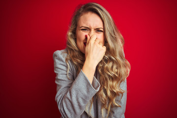 Young beautiful business woman wearing elegant jacket standing over red isolated background smelling something stinky and disgusting, intolerable smell, holding breath with fingers on nose.  - obrazy, fototapety, plakaty