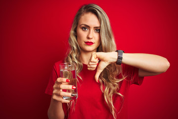 Fototapeta na wymiar Young beautiful woman drinking a glass of water over red isolated background with angry face, negative sign showing dislike with thumbs down, rejection concept