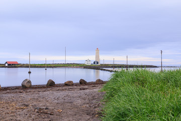 Coastline of the island of Grotta near Reykjavik with lighthouse, uninhabited houses and power pylons in the water. Blue sky with clouds and grass in the foreground