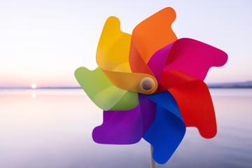 A round colored plastic pinwheel with a blowing wind stands on the sand by the sea against the smooth surface of the sea and a bright pink sunset. Toy mill on the sea beach. Large disk sun. Copyspace