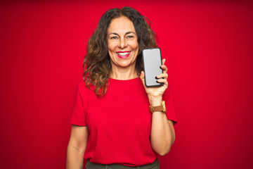 Middle age senior woman showing screen of smartphone over red isolated background with a happy face...