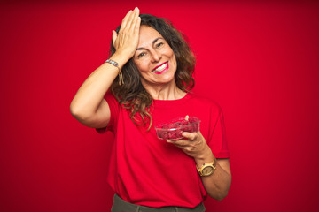 Middle age senior woman eating raspberries over red isolated background stressed with hand on head, shocked with shame and surprise face, angry and frustrated. Fear and upset for mistake.