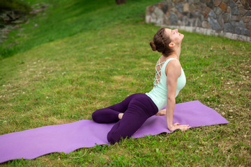 Slender young brunette yogi performs complex yoga exercise on green grass in the summer on a...