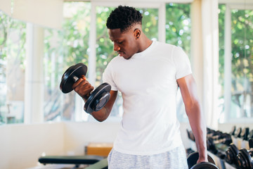 Young African American man standing and lifting a dumbbell with the rack at gym. Male weight...