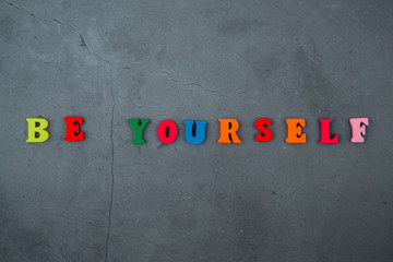 The multicolored be yourself word is made of wooden letters on a grey plastered wall background.