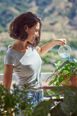 Brunette woman of Latin race waters the plants on a sunny terrace with views.