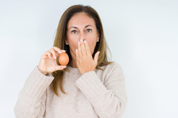 Middle age woman holding fresh egg over isolated background cover mouth with hand shocked with shame for mistake, expression of fear, scared in silence, secret concept