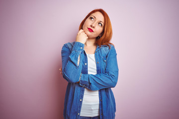 Youg beautiful redhead woman wearing denim shirt standing over isolated pink background with hand on chin thinking about question, pensive expression. Smiling with thoughtful face. Doubt concept.