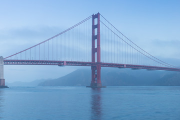 Fototapeta na wymiar Classic panoramic view of famous Golden Gate Bridge seen from San Francisco harbour in beautiful evening light on a dusk with blue sky and clouds and fog in summer, San Francisco, California, USA