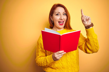 Young redhead teacher woman reading red book over yellow isolated background surprised with an idea or question pointing finger with happy face, number one