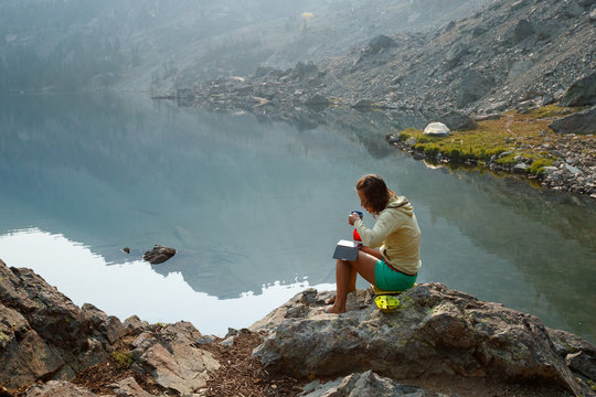 A female backpacker has her morning coffee next to Sheep Lake in the Seven Devils Mountains, Idaho, while reading from an electronic reader.