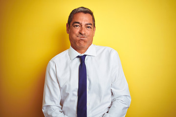 Handsome middle age businessman standing over isolated yellow background puffing cheeks with funny face. Mouth inflated with air, crazy expression.