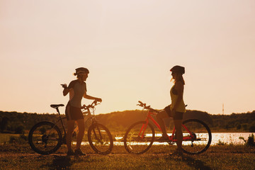 Two silhouettes of women with bike standing on lakeside and talking each other at the sunset