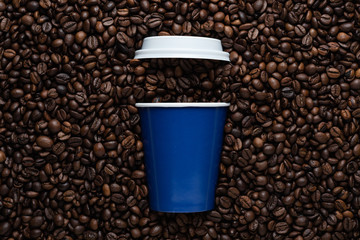 Blue disposable cup of coffee to go with a white lid on the background of roasted beans.