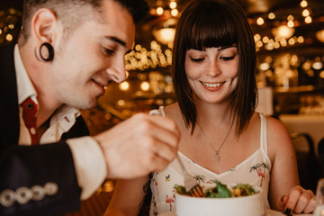Cheerful young man and woman eating tasty fresh salad while sitting at table on blurred background of luxury restaurant during romantic dinner