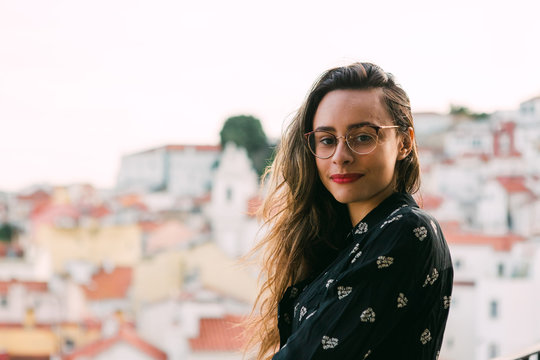 Portrait of smiling young brunette in glasses and with red lips looking at camera against blurred cityscape background