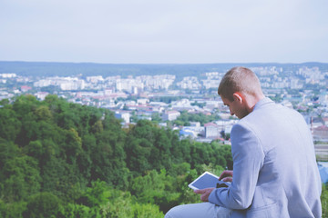 Concept: working anytime and anywhere. Young successful businessman working with digital tablet on top of the mountain. View from behind.