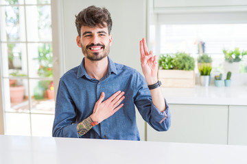 Young man wearing casual shirt sitting on white table Swearing with hand on chest and fingers,...
