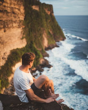Side view of man sitting alone on cliff at height looking at majestic landscape of ocean waves, Bali