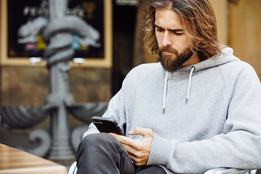 Young bearded handsome man in casual sweater sitting at cafe concentrated on mobile phone