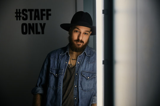 Young bearded handsome man in hat and denim shirt looking at camera in dark room