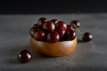 Red Cherry in a wooden cup on a gray background. some cherries are lying next to the plate. The background is dark