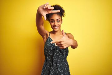 Naklejka premium African american woman wearing summer casual green dress over isolated yellow background smiling making frame with hands and fingers with happy face. Creativity and photography concept.