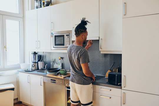 Side view of African American braided man standing in kitchen with frying pan and scapula on sunny day