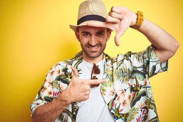 Young man on vacation wearing hawaiian flowers shirt and summer hat over yellow background smiling...