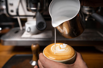 From above crop hands of professional employee preparing cappuccino with pattern on top in coffee shop