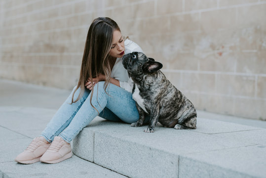 Casual young women sitting on concrete pavement kissing adorable bulldog on street