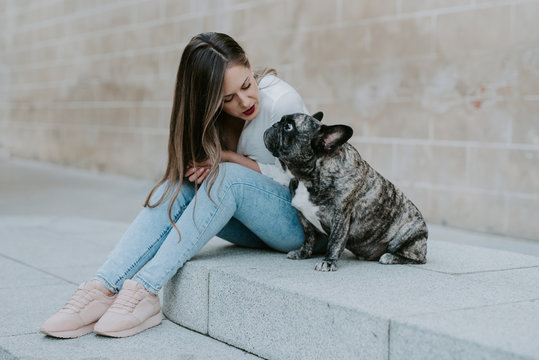 Casual young women sitting on concrete pavement kissing adorable bulldog on street