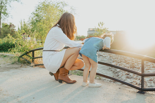 Side view of young woman and little girl bending over railing and looking at fast water stream on sunny day in park