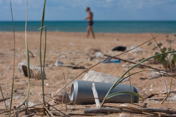 Fototapeta na wymiar Empty aluminum can and other garbage on the sea shore left by people, human figure walking on the background, Polluted dirty beach, Global ocean pollution