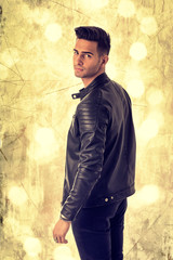 Young handsome man standing in studio shot, on golden background, wearing black leather jacket and black jeans