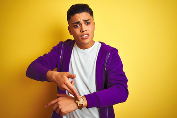 Young brazilian man wearing purple sweatshirt standing over isolated yellow background In hurry...