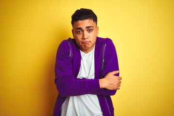 Young brazilian man wearing purple sweatshirt standing over isolated yellow background shaking and freezing for winter cold with sad and shock expression on face