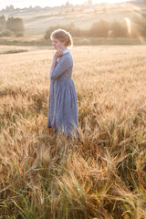 girl in blue dress standing at dawn in field of ears in golden light of sun. hill and country landscape in background