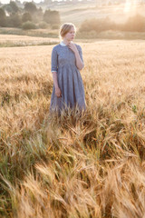 Naklejka na ściany i meble girl in blue dress standing at dawn in field of ears in golden light of sun. hill and country landscape in background