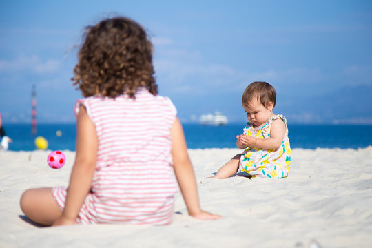 Baby girls in summer outfits sitting on beach and enjoying white sand on blurred sea background