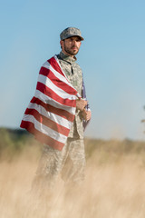 selective focus of man in camouflage uniform and cap holding american flag in golden field