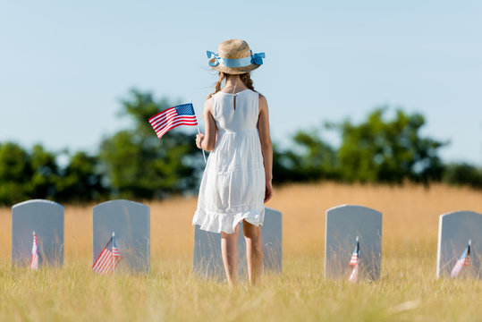 back view of child in dress and straw hat standing near headstone and holding american flag in graveyard