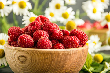 Raspberry berries in a wooden bowl on a dark table next to a bouquet of field camomiles. Summer background with garden berries and daisies. Delicious red ripe berries in a wooden bowl.